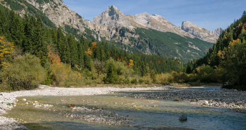 The wide Karwendelbach river running through the Karwendel Valley. In the background are alpine peaks with woodland reaching high up the sides of the mountains. The Adlerweg route runs down this valley. 