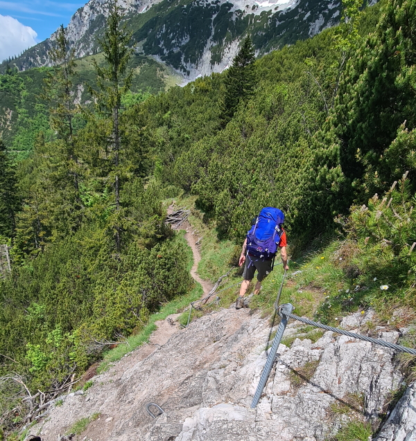 A hiker uses a fixed chain to aid with corssing a rocky section of path on the Adlerweg trek through the Austrian Tyrol. In the background the path continues into trees.