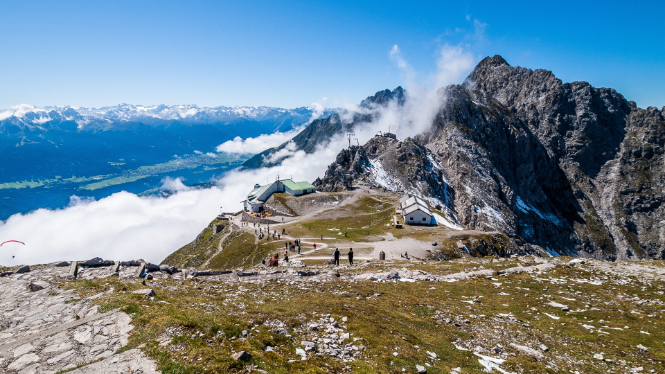 View from the Goetheweg optional day hike. The view is of the Nordkette cable car station with rocky mountains behind. Low wispy clouds partially cover the mountains and below the view of the Inn Valley is revealed