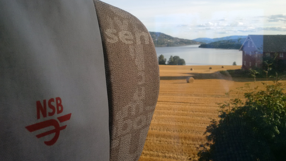 A Norwegian train headrest as the train passes a field and a barn. This is the old NSB logo, now Vy.